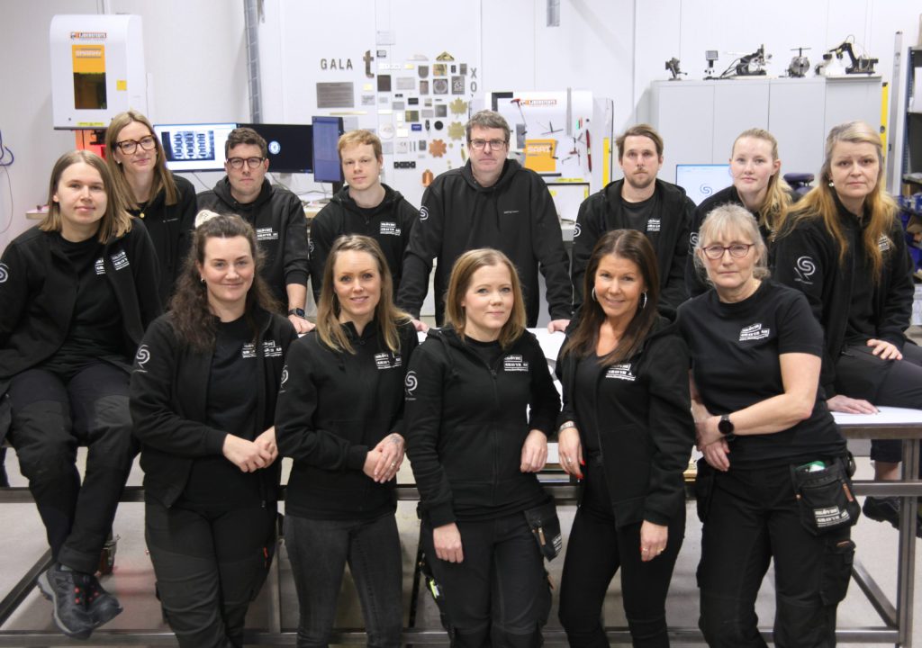 Skövde gravyrs medarbetare samlade i en gruppbild i industrilokal, iklädda svarta företagskläder med leenden på läpparna.